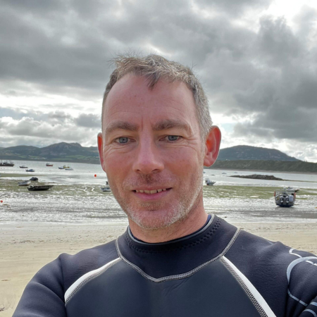 David King is standing on the beach wearing a wetsuit. The clouds in the background are dark.